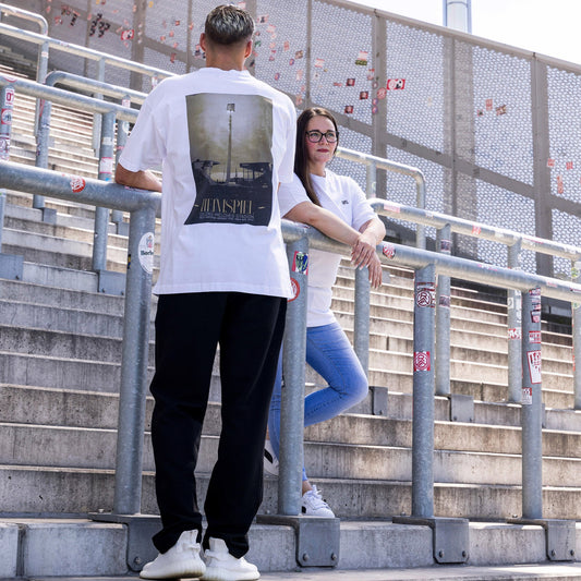 Jasmin und Marek tragen weißes oversize T-Shirt mit großem Fotoprint des alten Flutlichtmasts vor dem Georg-Melches-Stadion in braun schwarz gehalten; vorne "GMS" Stick in gold