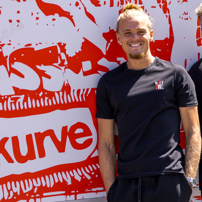 Tom Moustier trägt schwarzes Regular Fit T-Shirt mit rot-weiß-grauer "NUR DER RWE"-Stickerei auf Herzseite und rotem Nackenband