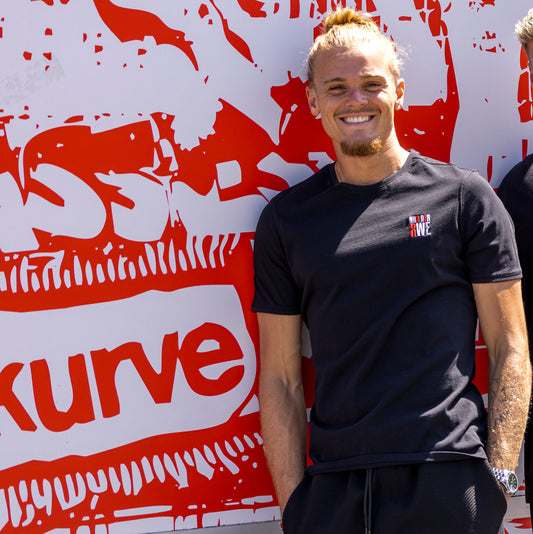 Tom Moustier trägt schwarzes Regular Fit T-Shirt mit rot-weiß-grauer "NUR DER RWE"-Stickerei auf Herzseite und rotem Nackenband