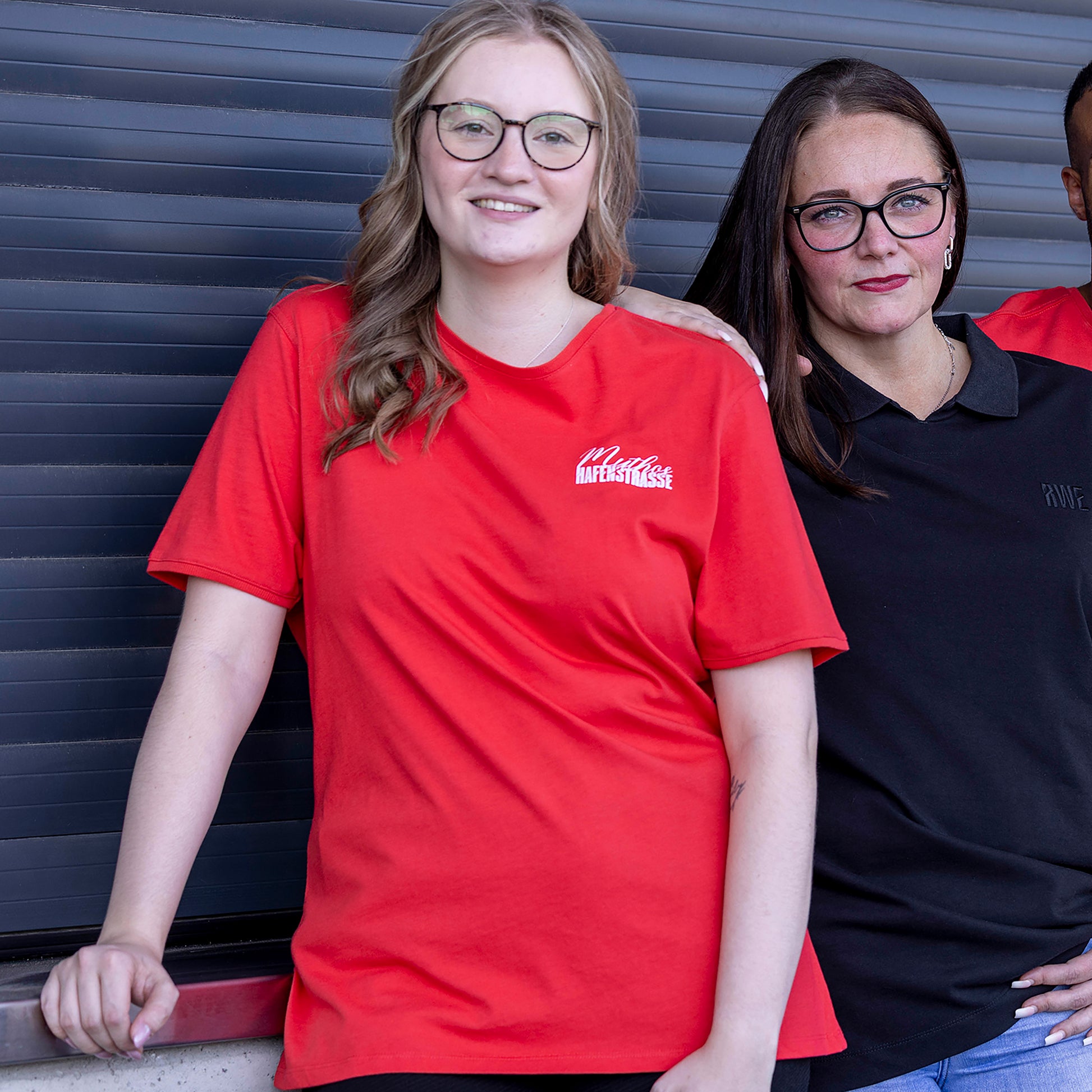 Marie trägt rotes T-Shirt im Regular Fit mit einem weißen "Mythos Hafenstrasse" -Druck auf der linken Brust. Der Druck ist eine Mischung aus Kalligraphie-Schrift (Mythos) und Rot-Weiss Essen-Schrifttyp (blockig; Hafenstrasse). Mit Rippbündchen am Ausschnitt und Ärmeln.