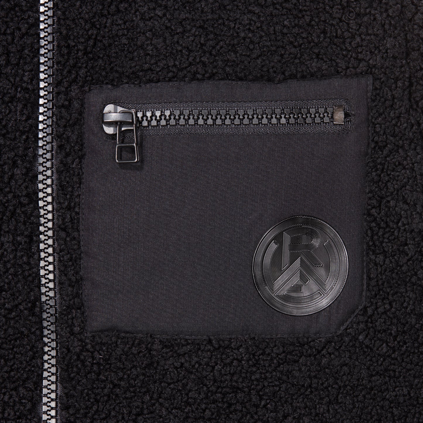 Nahaufnahme Brusttasche aus glattem Kontraststoff, mit Reißverschluss und Silikon Logo-Patch Ton-in-Ton zum Teddystoff