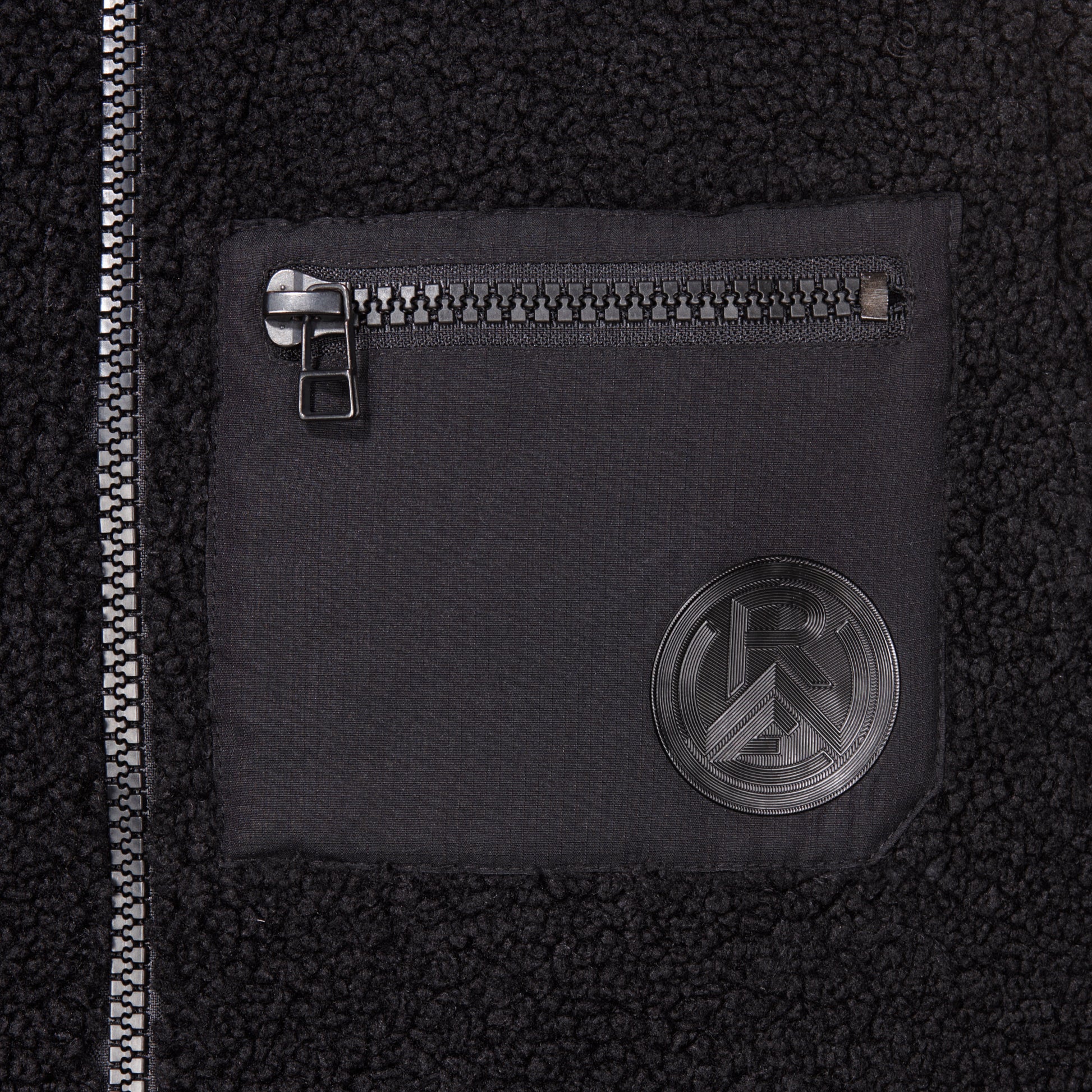 Nahaufnahme Brusttasche aus glattem Kontraststoff, mit Reißverschluss und Silikon Logo-Patch Ton-in-Ton zum Teddystoff